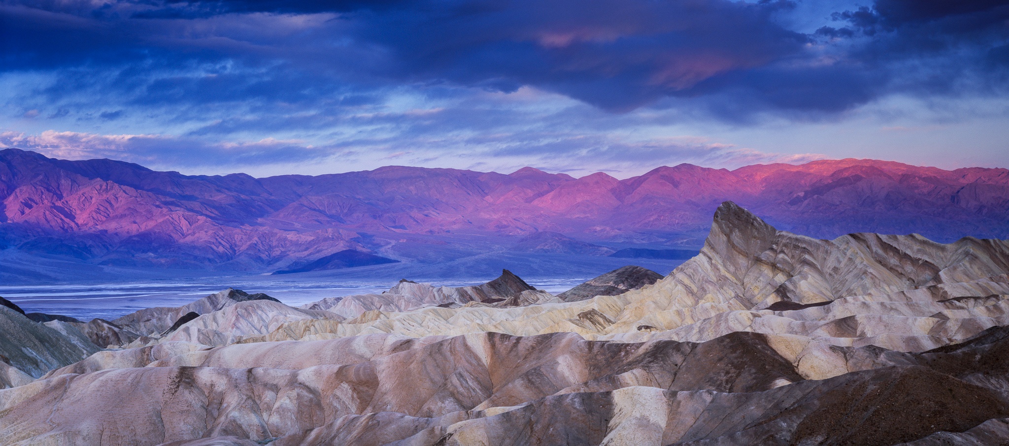 Pre-dawn light at Zabriskie Point, the eroded badlands pale and sculptural in the foreground while alpenglow catches the Panamint Range across the valley, California. Photograph by Marty Quinn