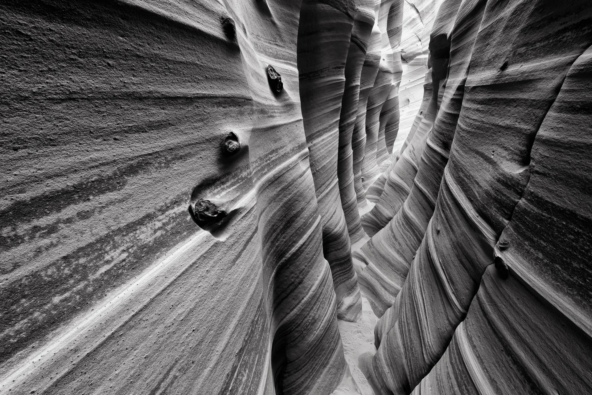 Diagonal bands of cross-bedded Navajo sandstone line the walls of a Utah slot canyon in this black and white study of layered rock and narrow light. Photograph by Marty Quinn